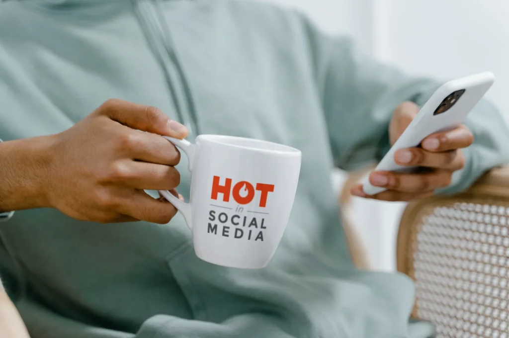 A man wearing a green sweater sits on his phone, holding a white mug with the Hot in Social Media logo