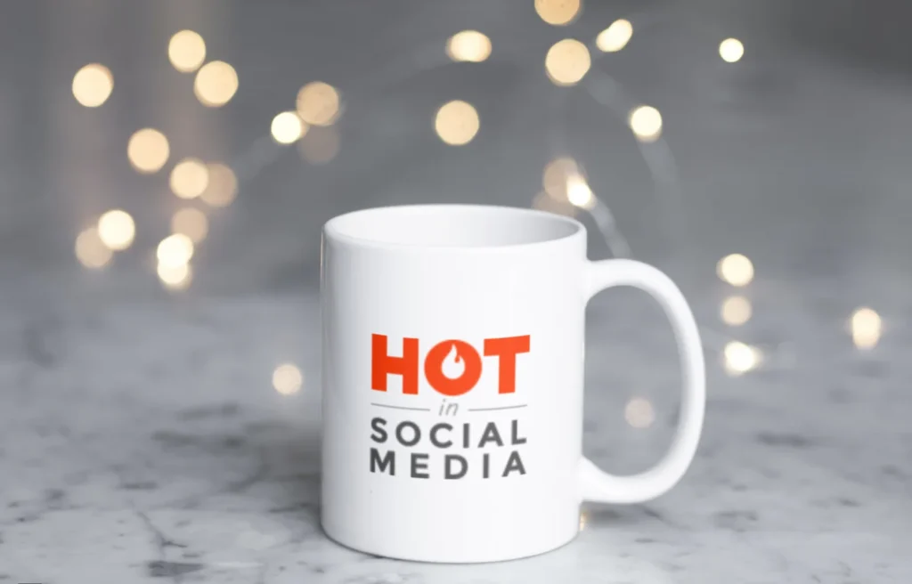 The Hot in Social Media logo on a white mug on a marble table with twinkle lights in the background